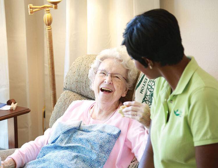 Caregiver leans over, speaking to her smiling senior client