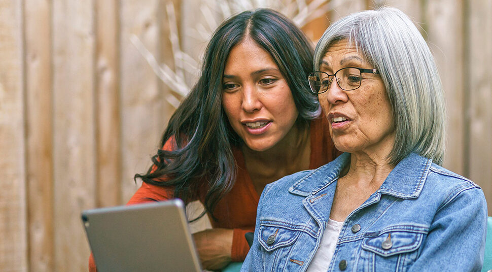 A senior reads information on a laptop, while a caregiver looks over her shoulder