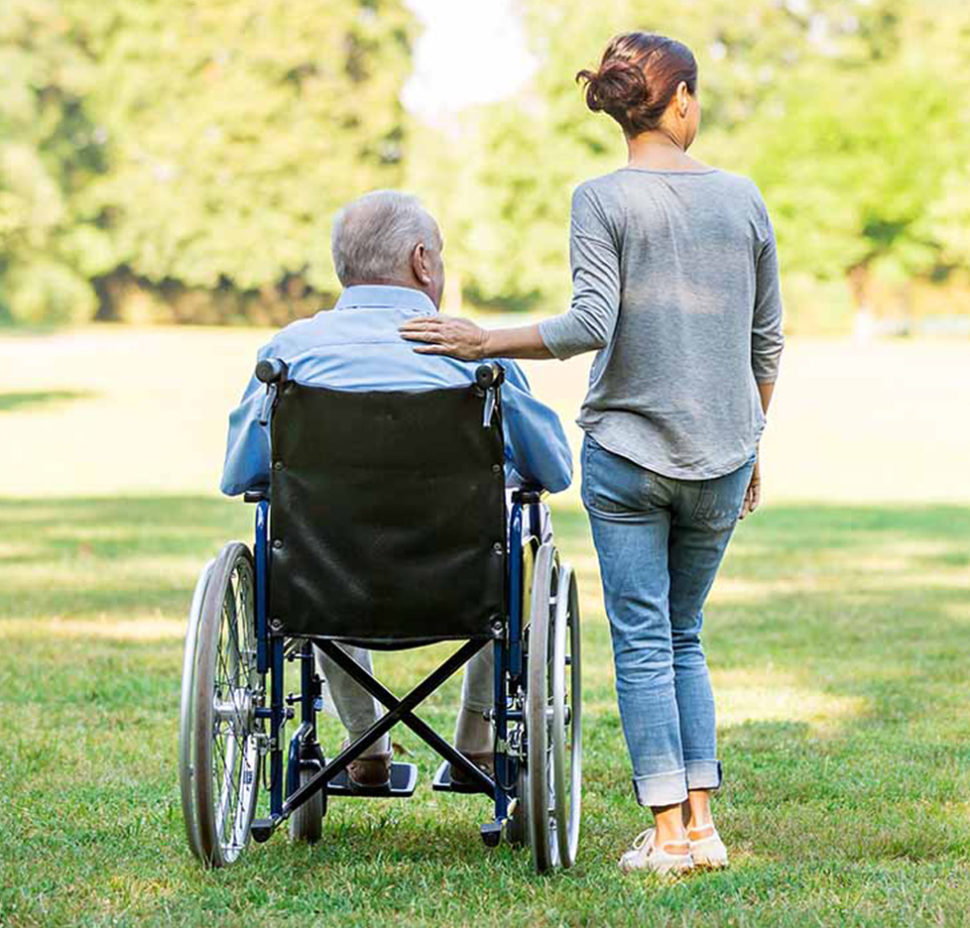 A caregiver stands beside an elderly man in a wheelchair, outdoors