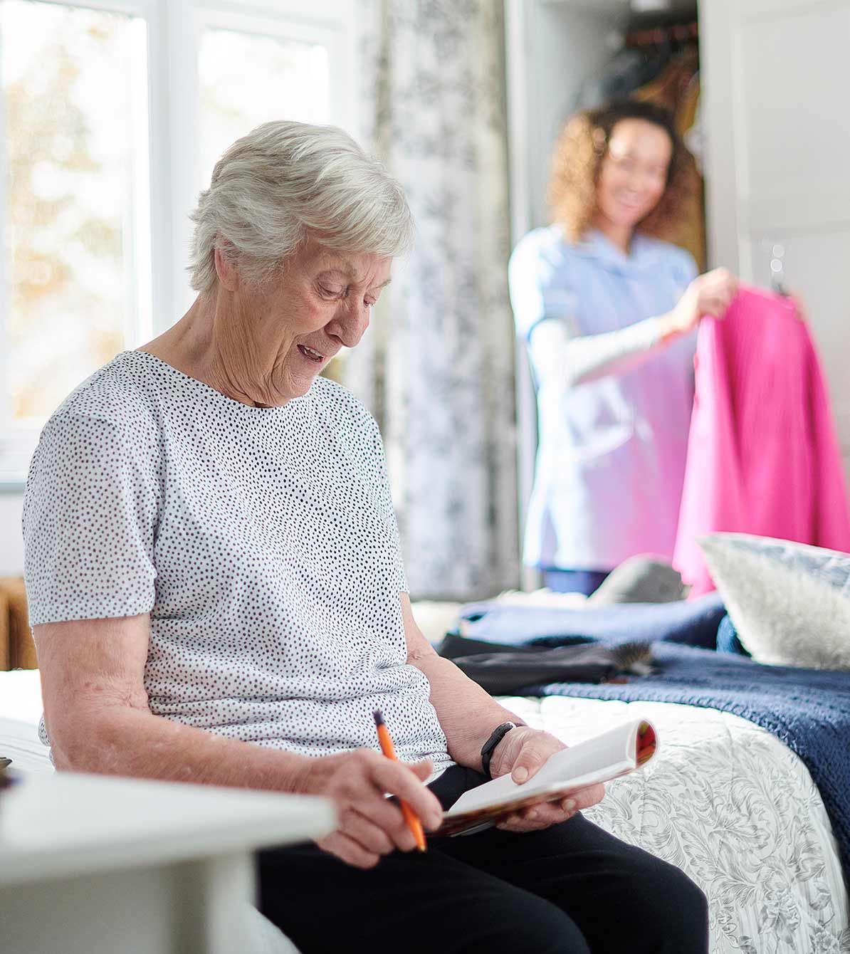 A caregiver folds laundry, while her client reads a magazine