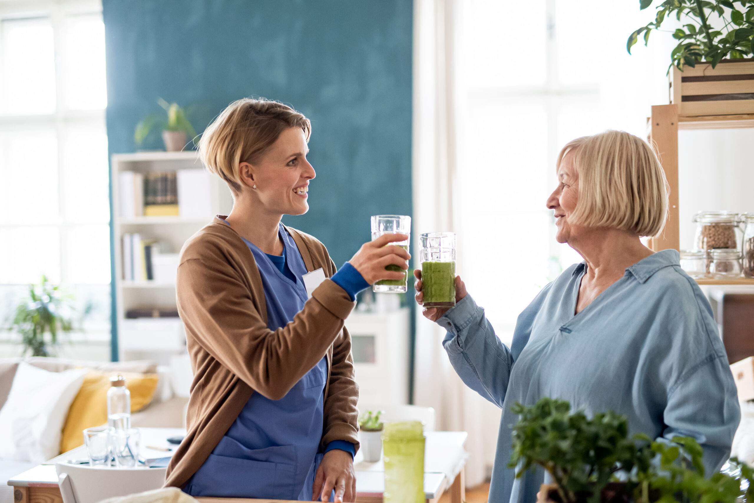 Two women smile and toast with green drinks in a bright living room.