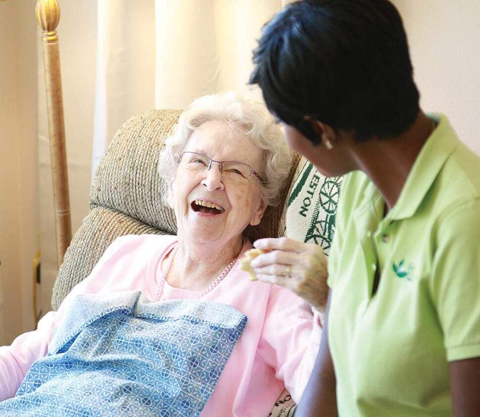 Caregiver leans over, speaking to her smiling senior client