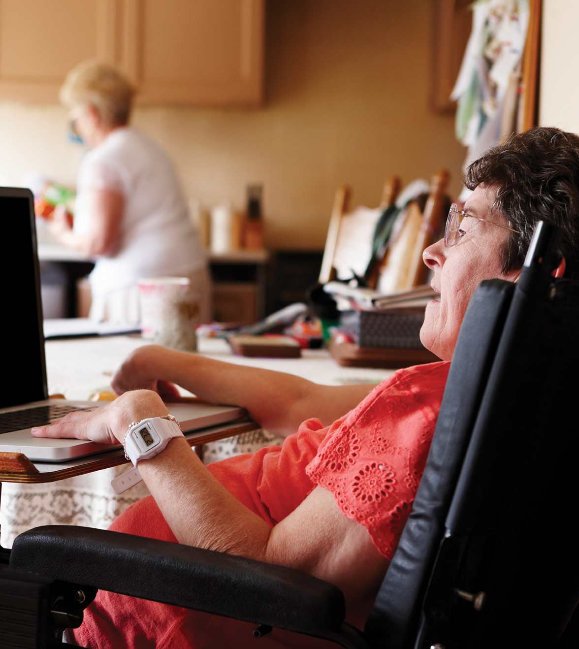 A woman in a wheelchair sits at her computer, while her caregiver makes food in the kitchen