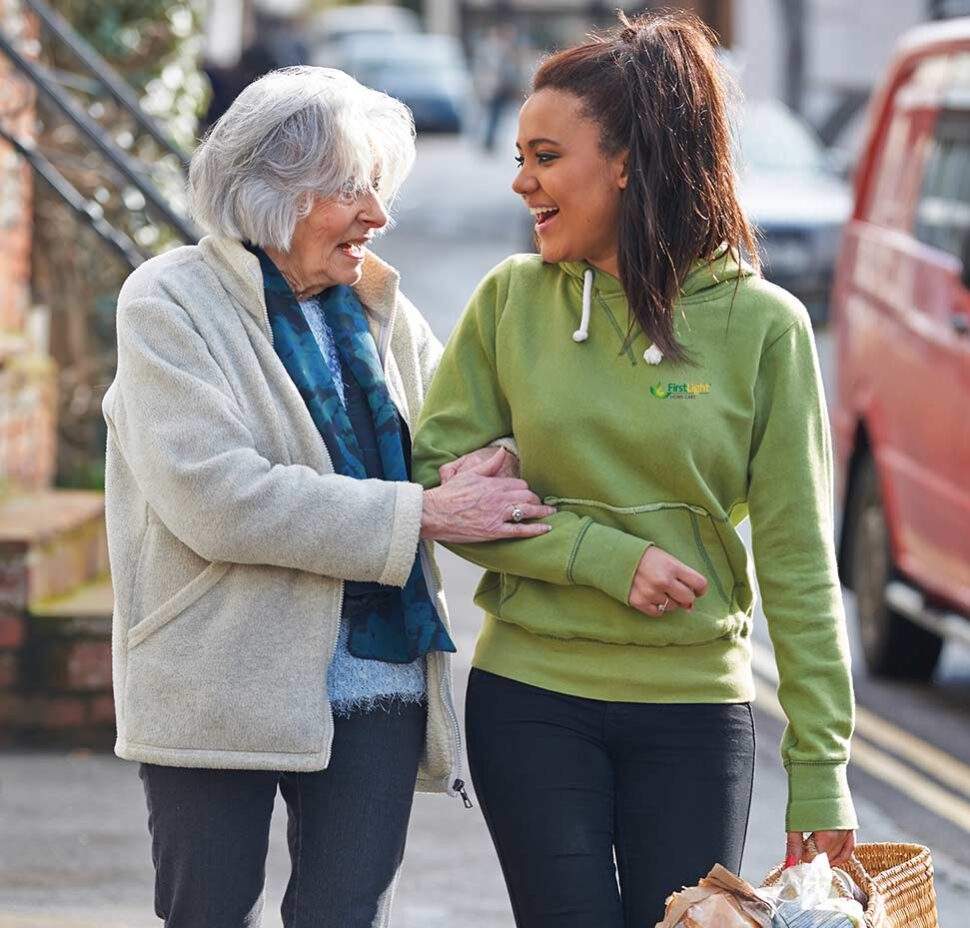 A FirstLight caregiver walks with a senior client. The caregiver is holding the client's bag and providing an arm for support