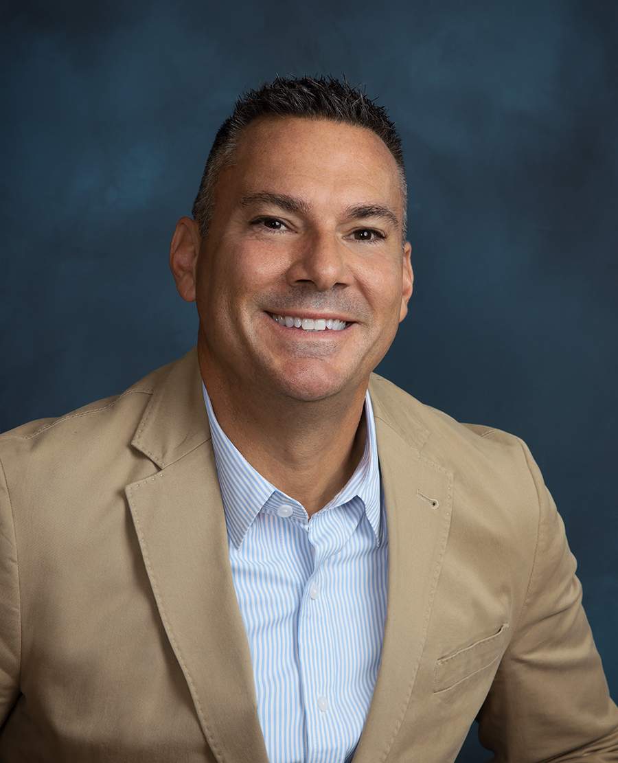 Smiling man in a tan blazer and striped shirt against a dark blue background.