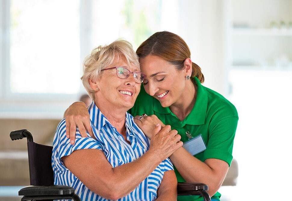 An older woman in a wheelchair smiles as her caregiver gives her a hug.