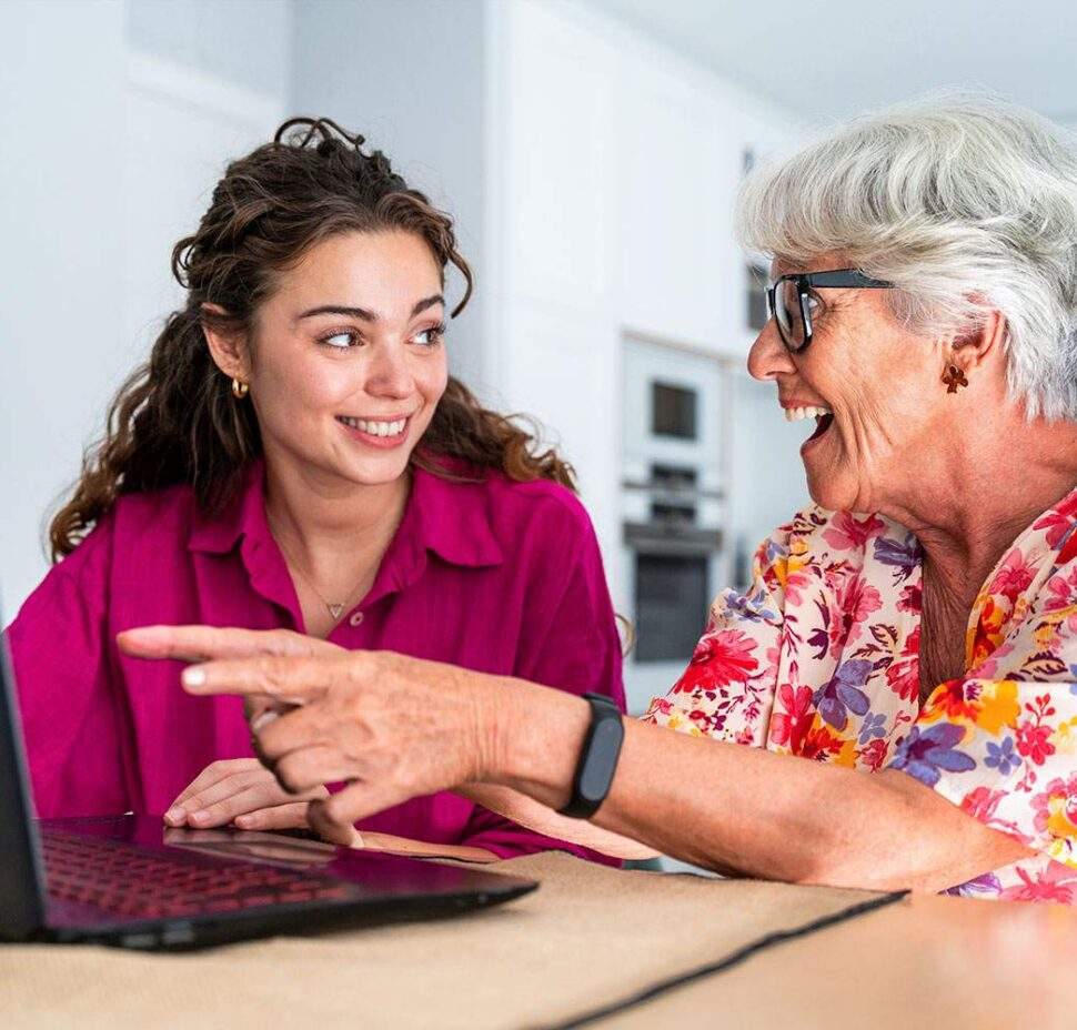 A personal care attendant smiles as an older woman shows her something on her laptop.