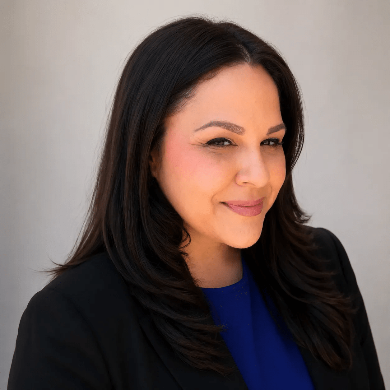 A smiling woman with dark hair wearing a black blazer and blue top against a light background.