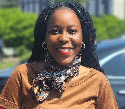 Smiling woman outdoors in front of a car, wearing a patterned scarf and brown top.