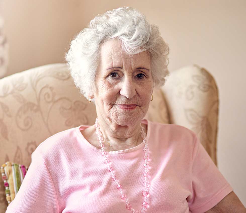 A senior woman in a pink shirt leans back in a large stuffed chair in her home