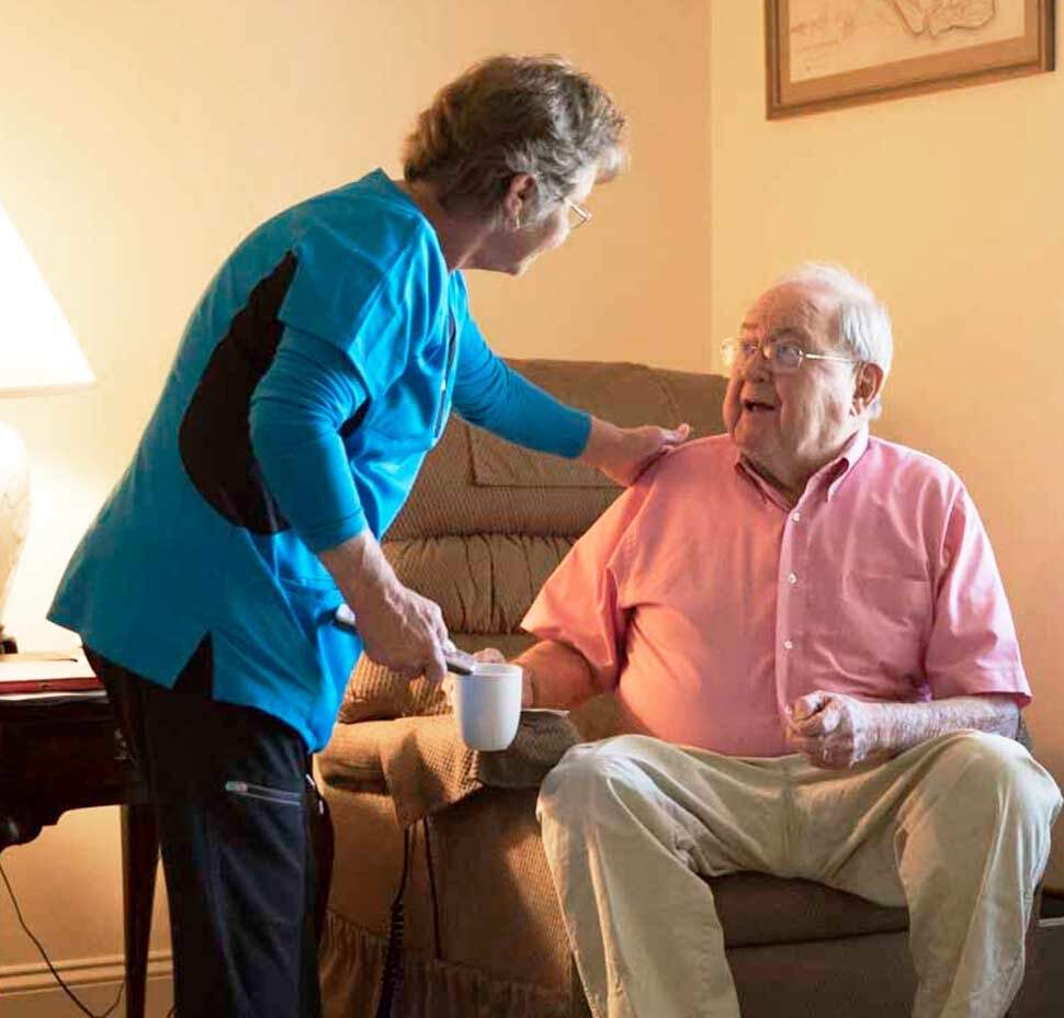 A caregiver leans over to speak with a client who is sitting in a chair in his living room