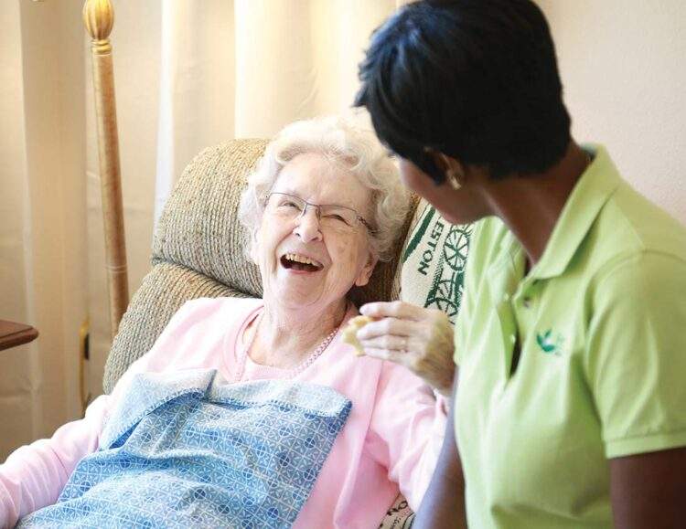 Caregiver leans over, speaking to her smiling senior client