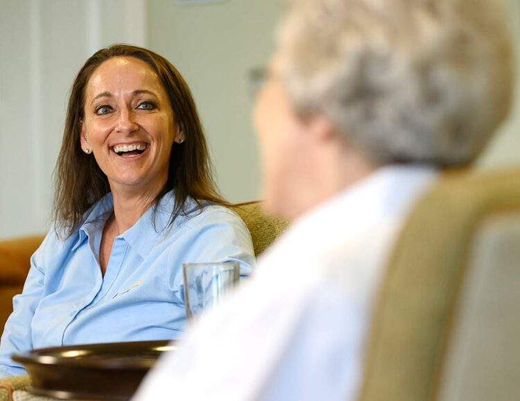 A caregiver sits across from her client, a senior woman, and smiles at her