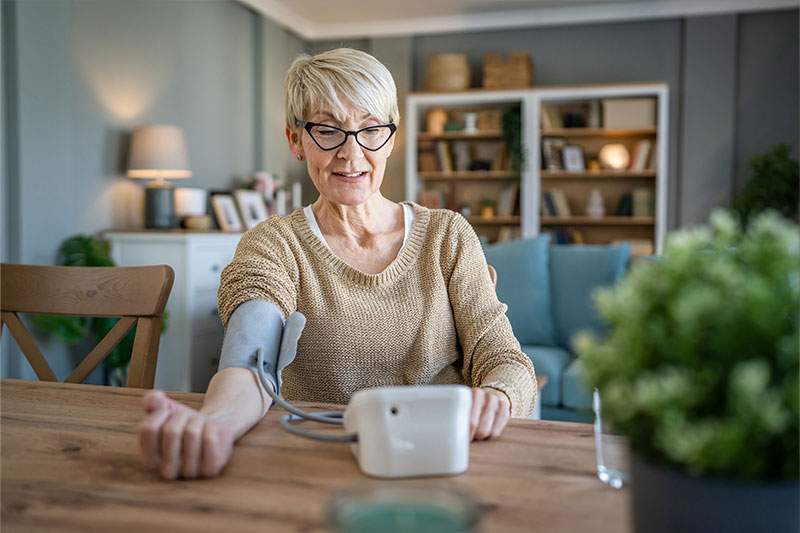 A woman with glasses sits at a table, using her blood pressure monitor for health monitoring.